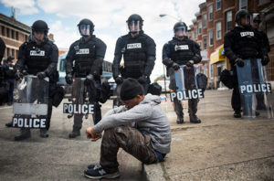 Baltimore, 2015: Daquan Green, age 17, sits on the curb while riot police stand guard after the funeral of Freddie Gray.(Photo by Andrew Burton/Getty Images)